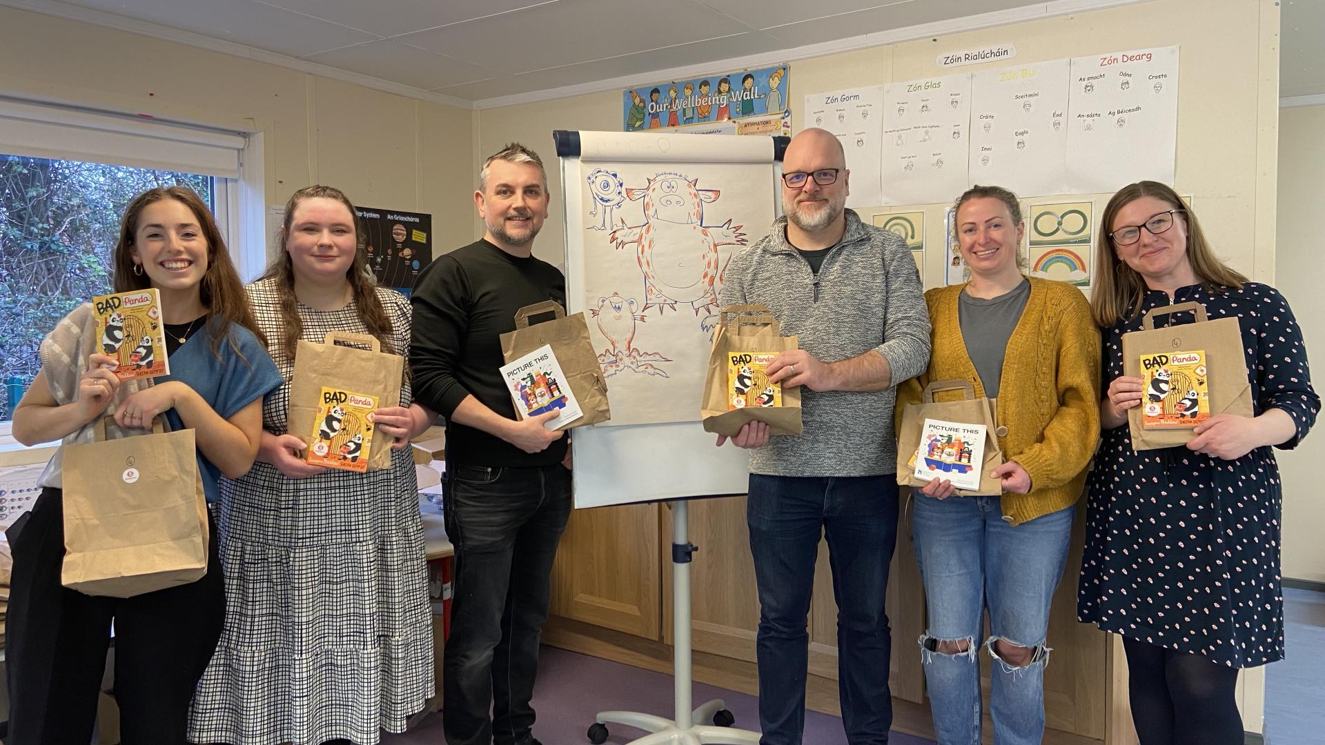 Group holding Bookbag packages and children’s books in a classroom beside a flip chart with character sketches.