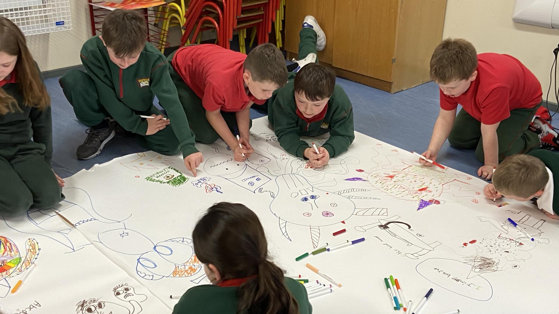 Children lying on the floor drawing colorful characters on a large sheet of paper.