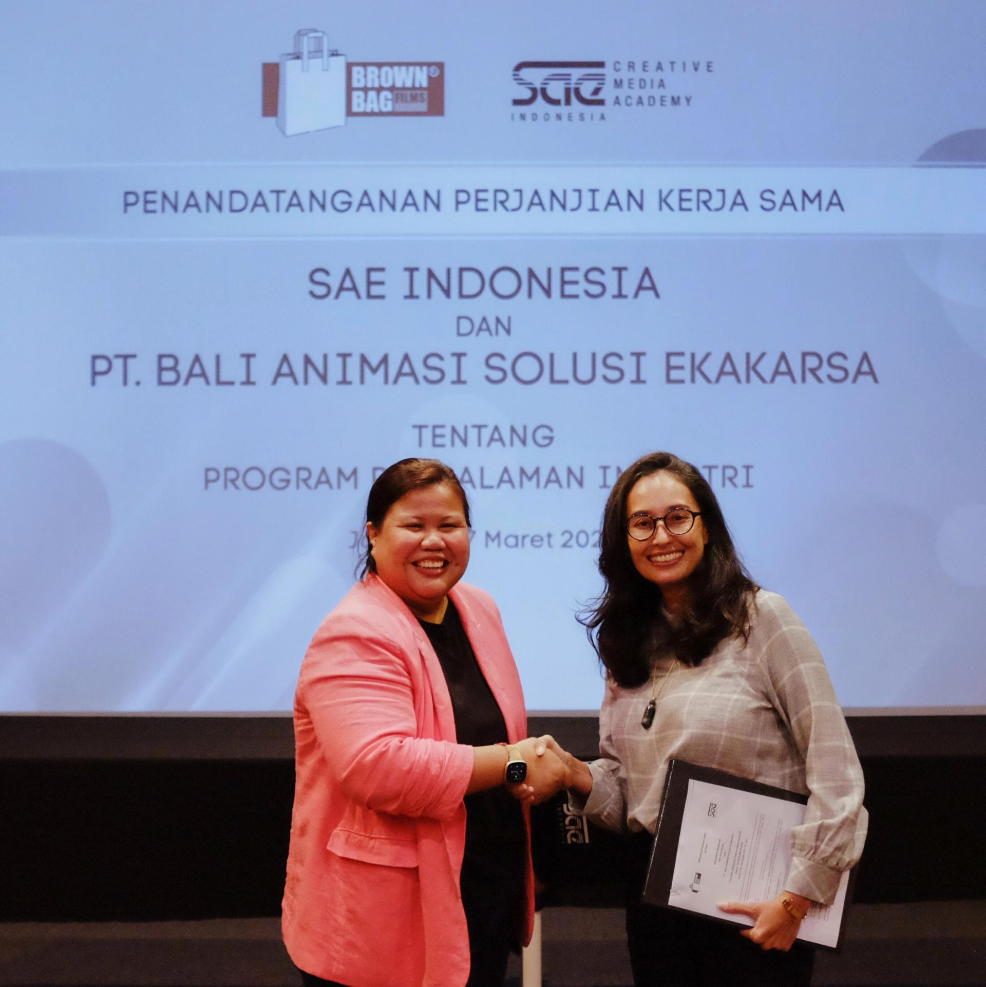 Two people shaking hands in front of a presentation screen displaying partnership details between SAE Indonesia and PT. Bali Animasi Solusi Ekakarsa.