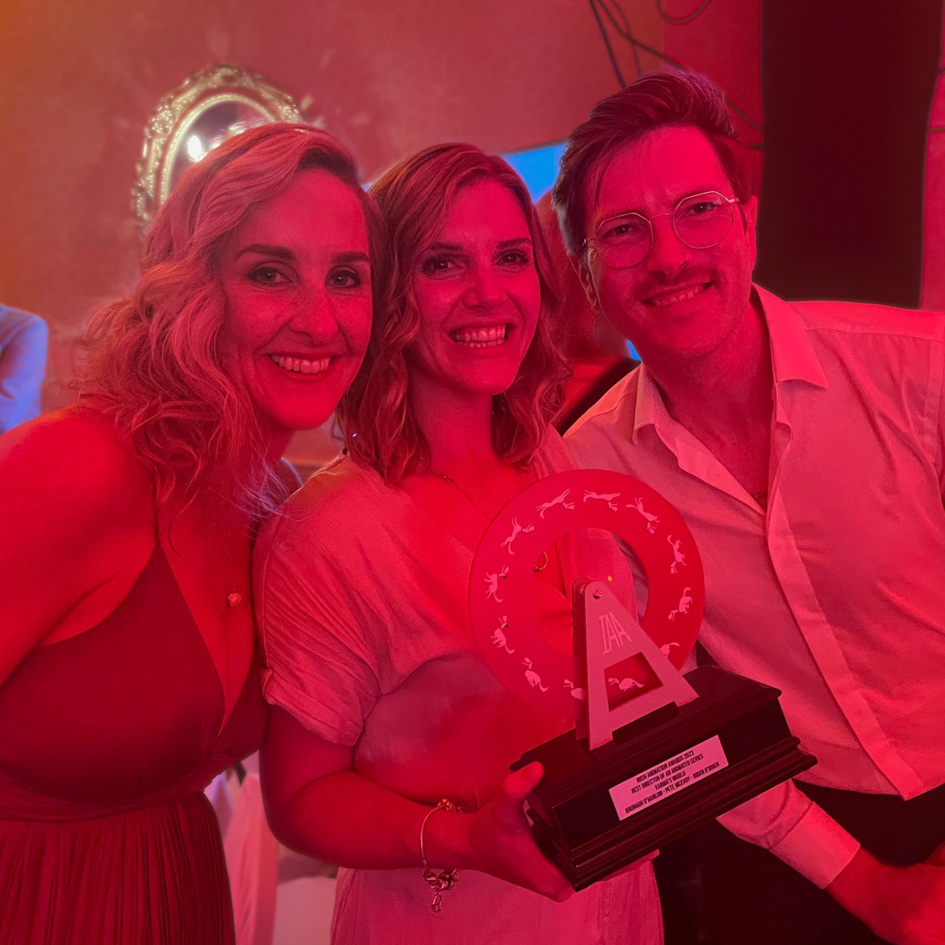 Three people posing together holding Irish Animation Award trophy under red lighting.