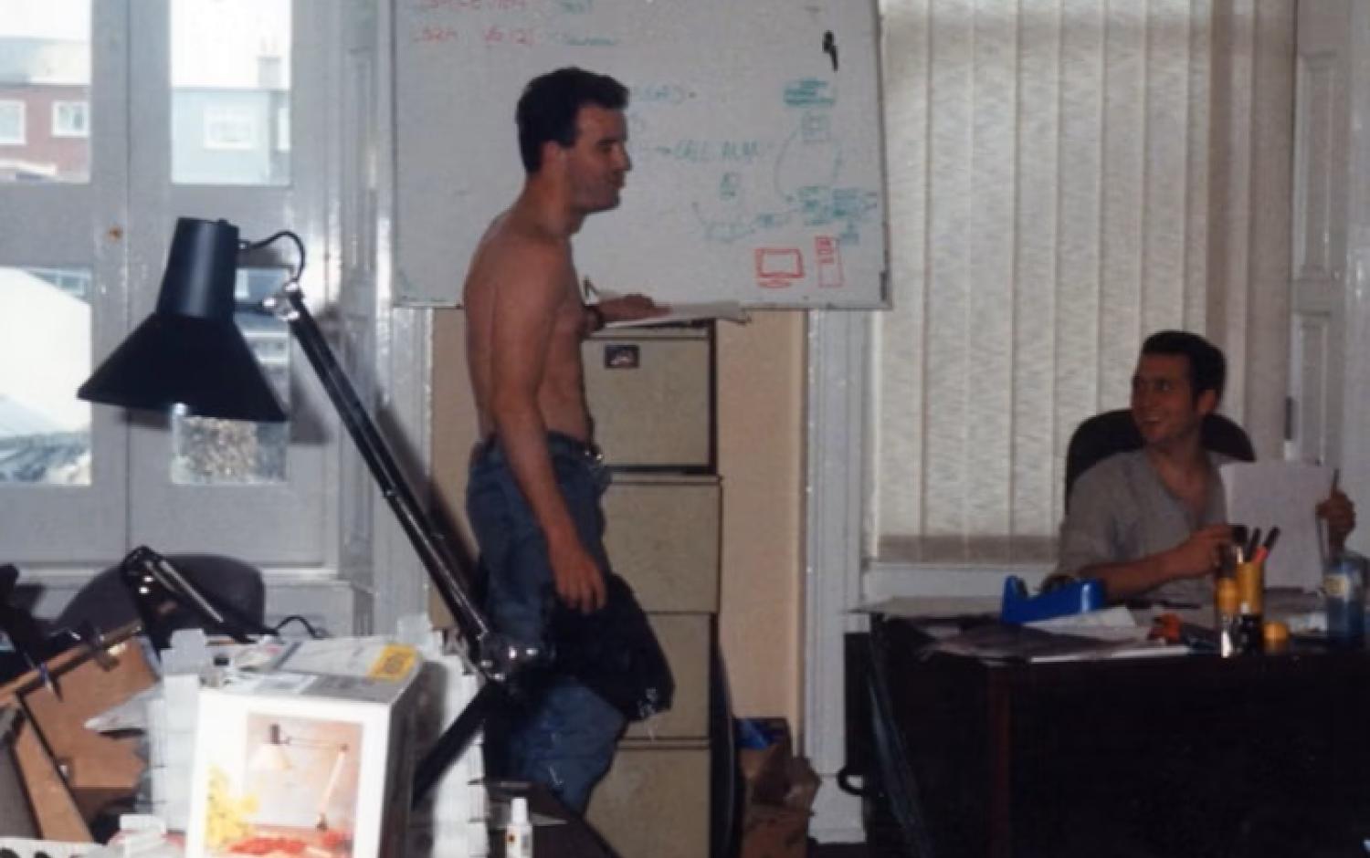 One person standing by a desk with computer equipment, another seated nearby, both working.
