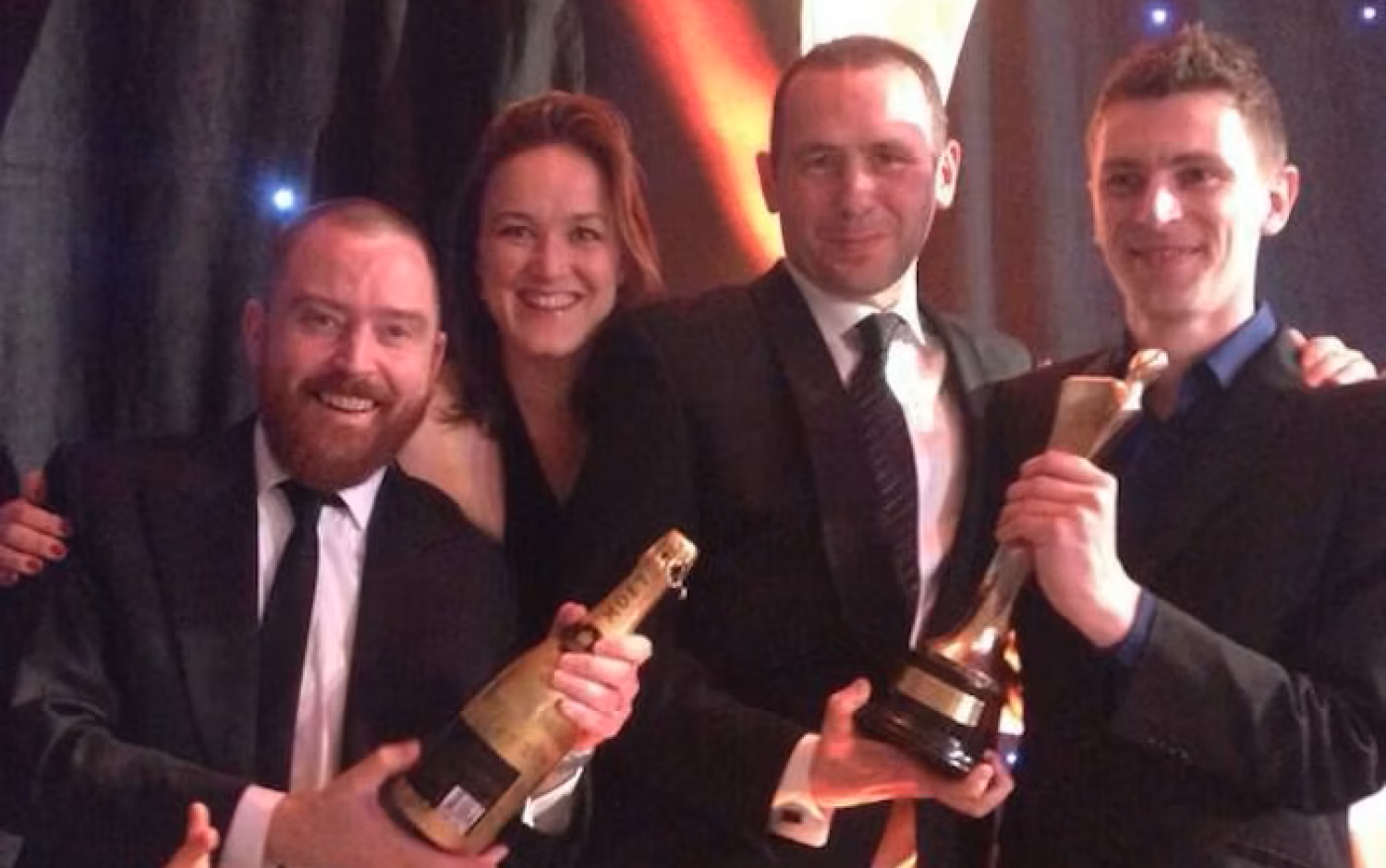 Four people in formal attire smiling and holding awards and champagne bottles in front of a spiral-themed backdrop, while someone takes their photo with a smartphone.