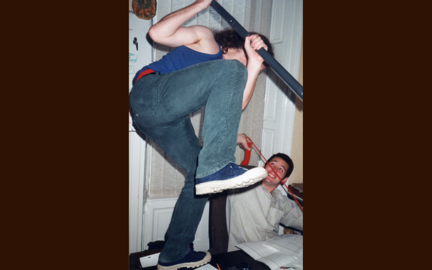 Two people in an office engaged in a playful mock fight; one person is standing on a desk holding a large metal pole, while the other sits below holding a red phone.