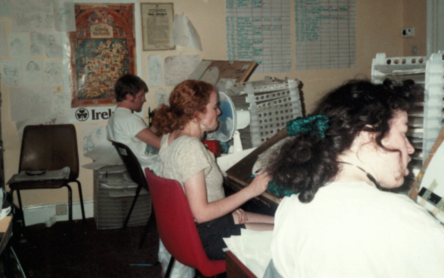 Three animators seated at desks painting animation cels, surrounded by shelves and pinned artwork.