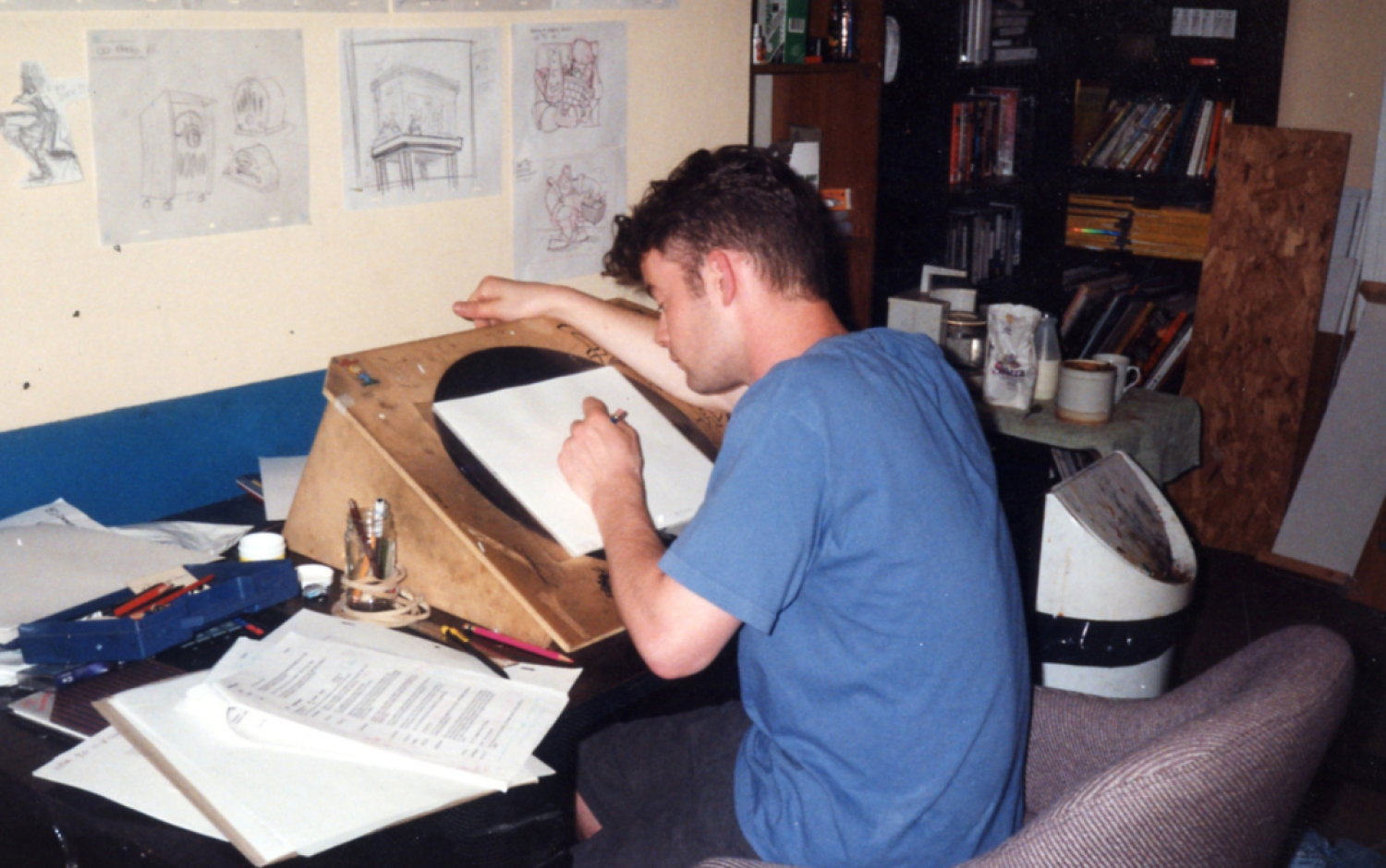Animator working at a wooden animation desk, sketching on paper; character drawings are pinned to the wall behind.