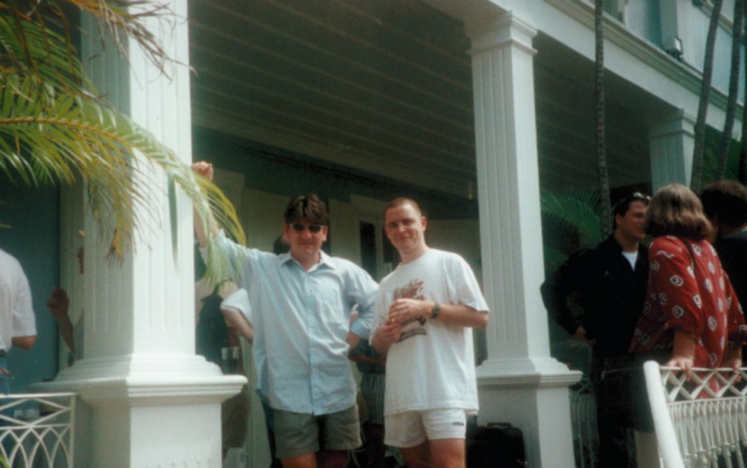 Group of people gathered on a porch with white columns and tropical plants, two individuals standing in the foreground.