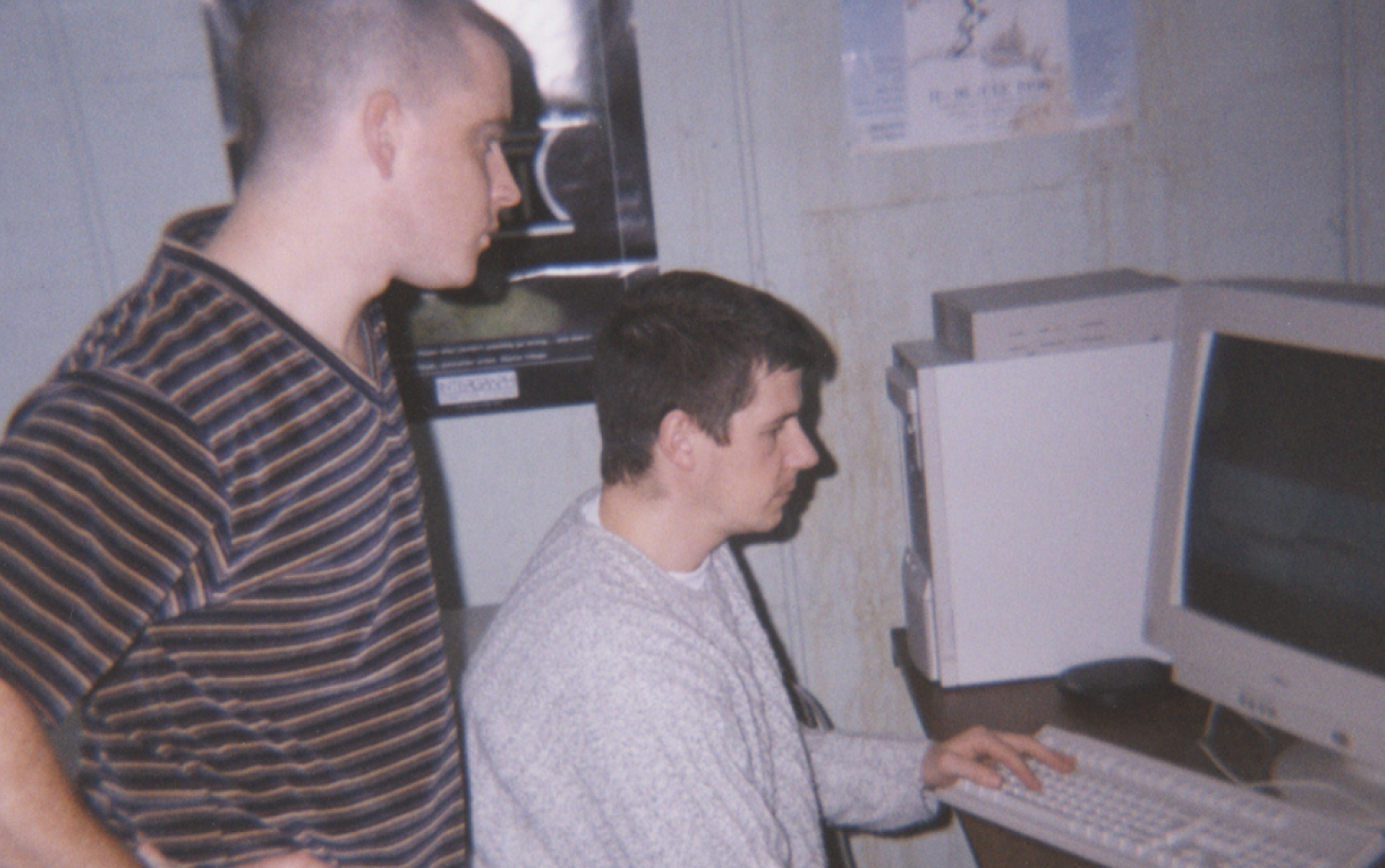 Two people in a small office setting working at a desk with a CRT monitor and keyboard; one person is seated using the computer while the other stands nearby observing.