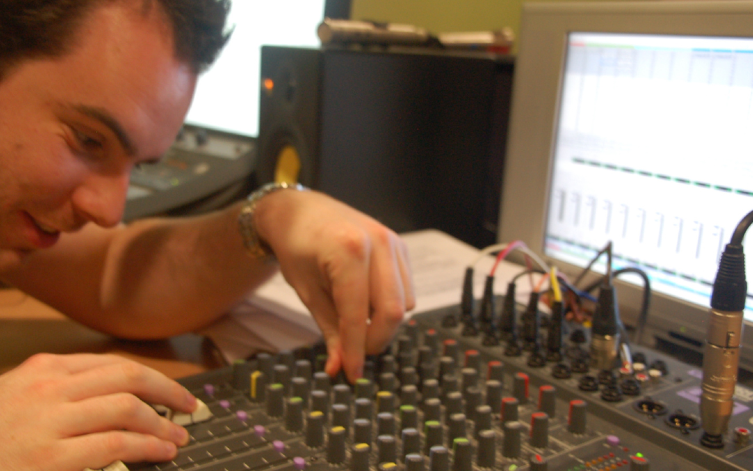 Close-up of hands adjusting knobs on an audio mixing console with a computer screen displaying sound levels in the background.
