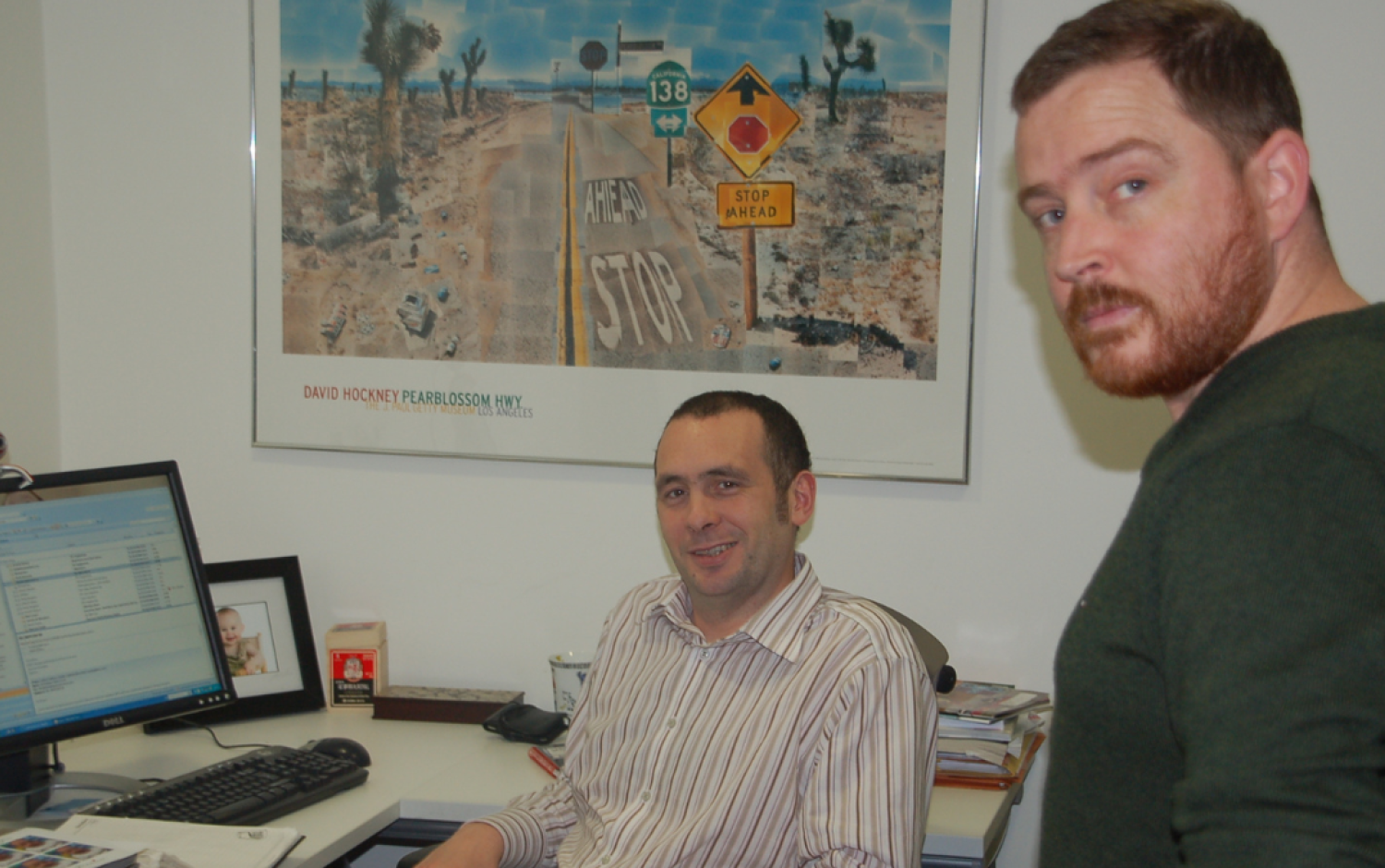 Office scene with two people at a desk, one seated at a computer displaying an email interface, and a large framed artwork on the wall.
