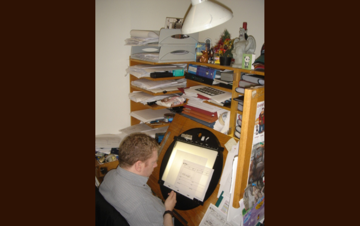 Animator working at a lightbox in a small, cluttered workspace with shelves of papers and office supplies.