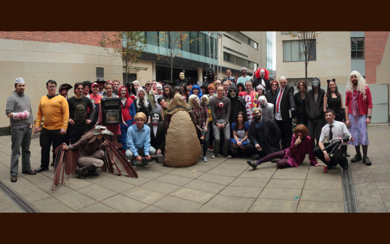 Large group of people in costumes gathered in a courtyard for a Halloween event, featuring various props and themed outfits.