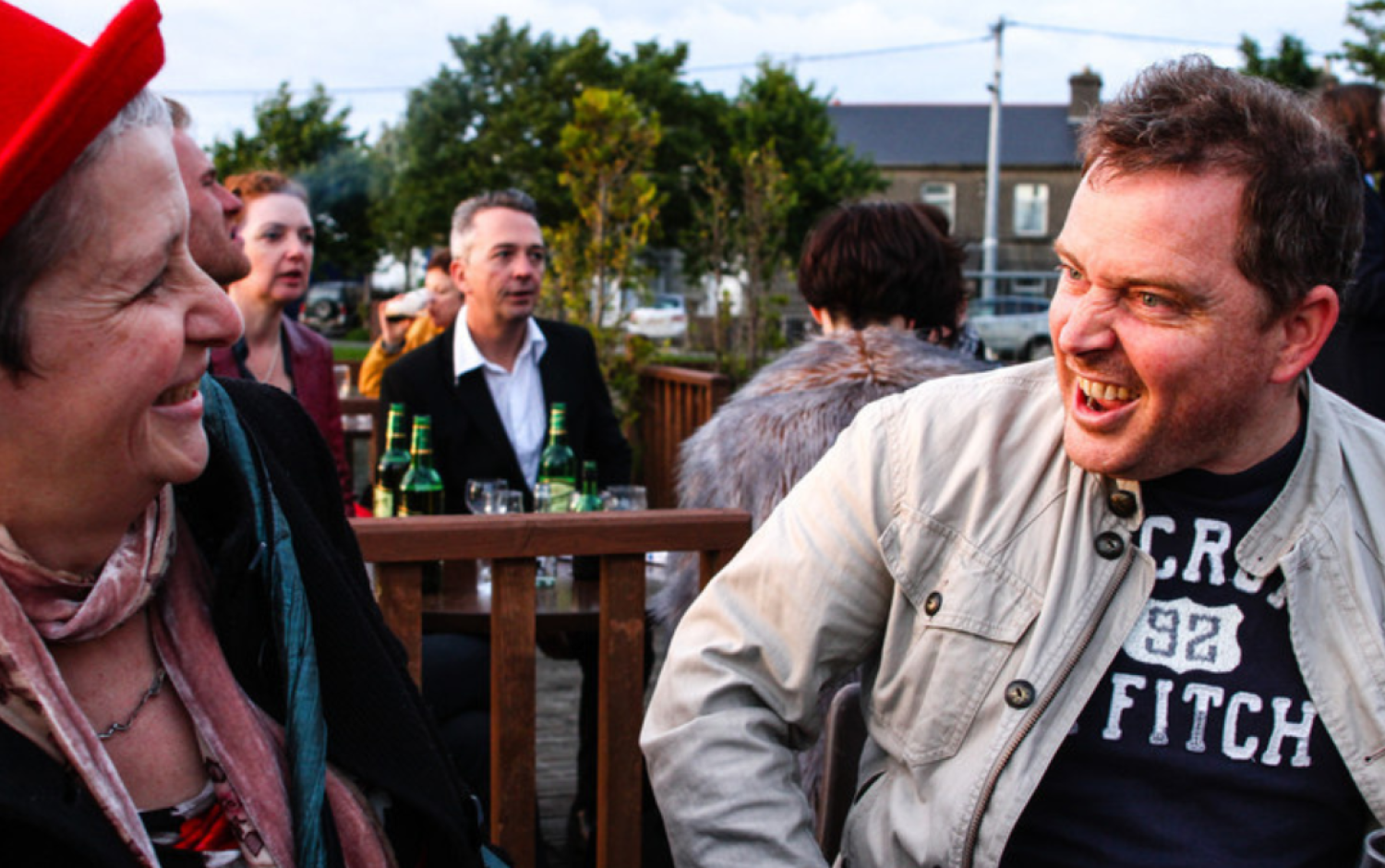 Two people seated outdoors at a wooden table, talking; several bottles and glasses are visible on tables in the background.