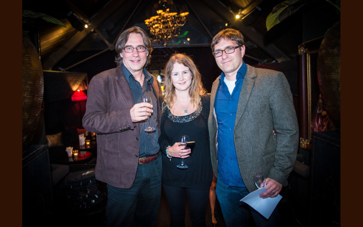 Three people standing together in a dimly lit venue holding wine glasses, with a chandelier overhead.