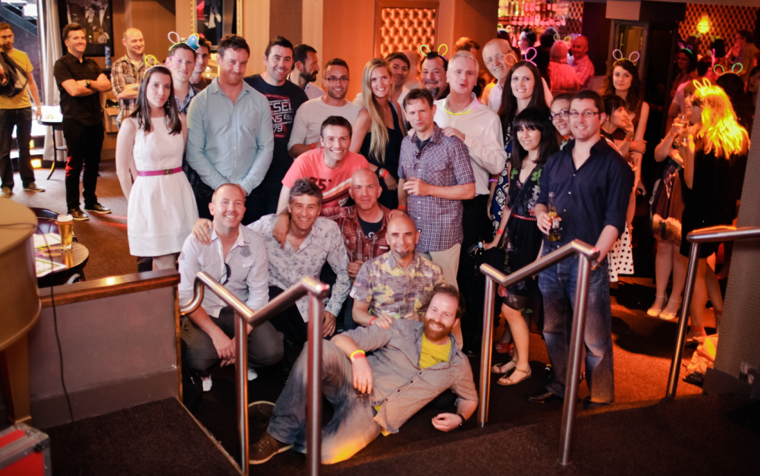 Large group gathered in a bar or club setting, some standing and others seated on steps, with drinks in hand.