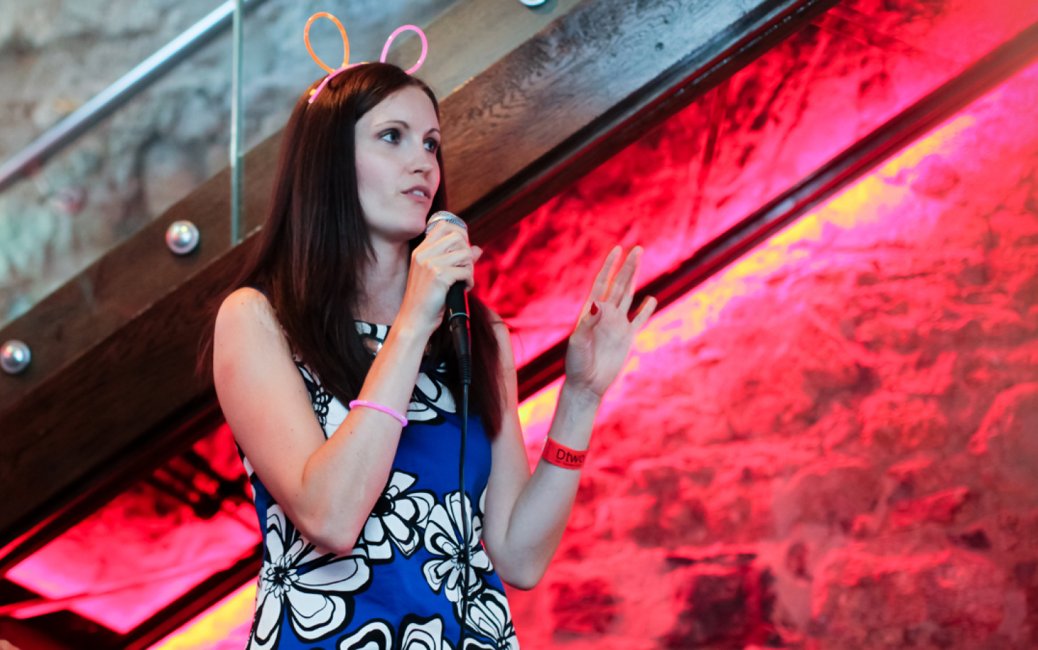 Person speaking into a microphone on a stage with red lighting and a wooden staircase in the background.