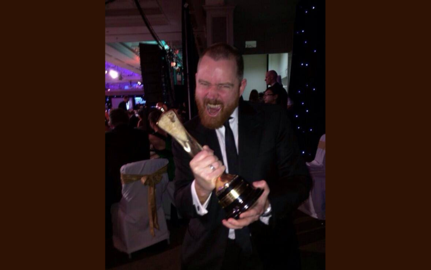 Person in formal attire holding a tall award trophy at an event with decorated tables.