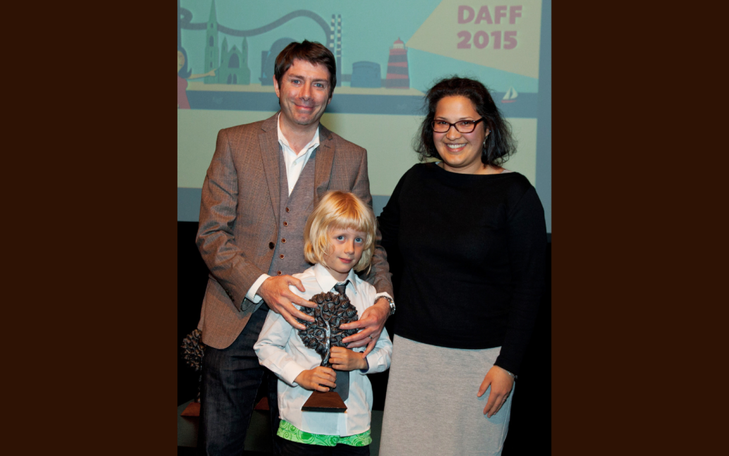 Three people posing on stage at DAFF 2015, child holding a tree-shaped award.