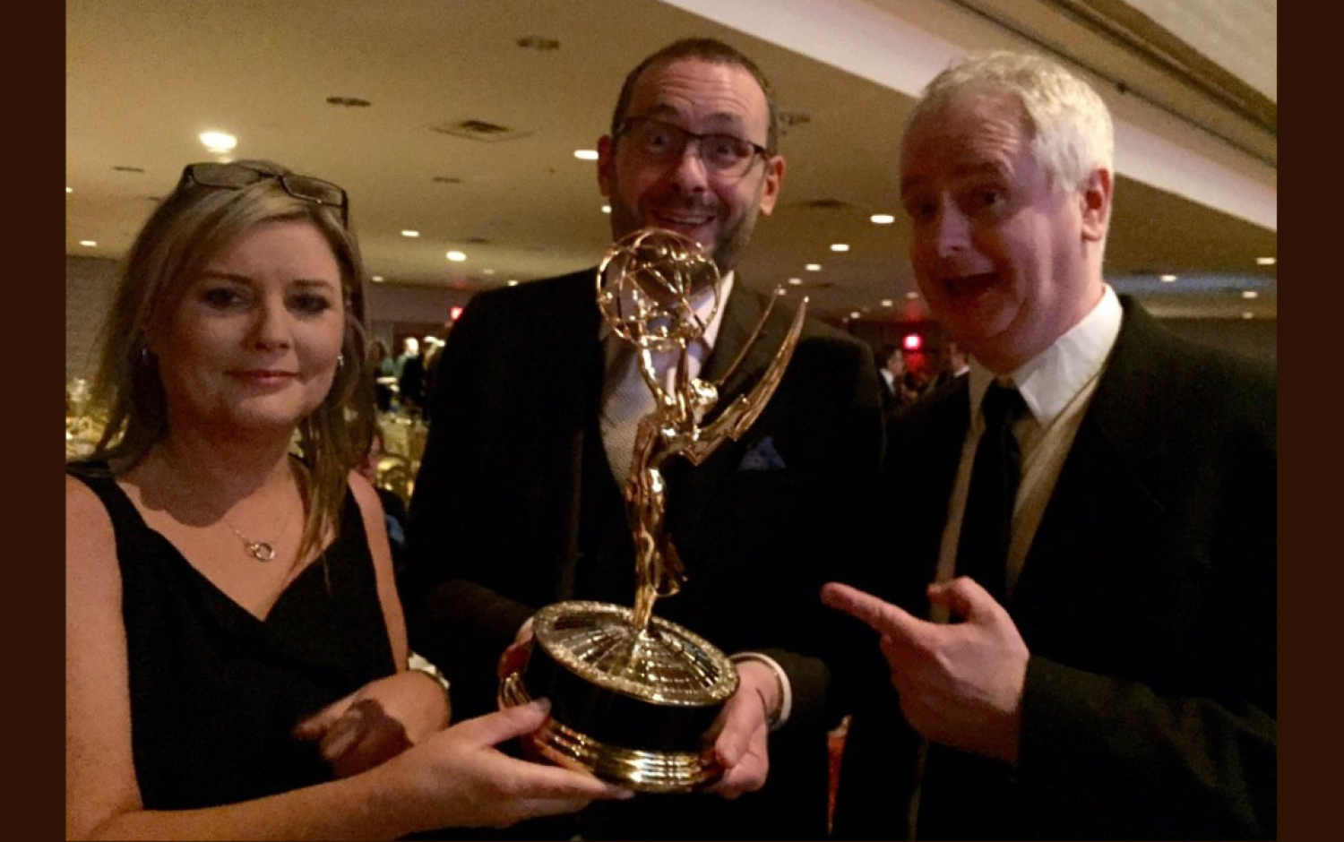 Three people holding an Emmy Award trophy at a formal event.