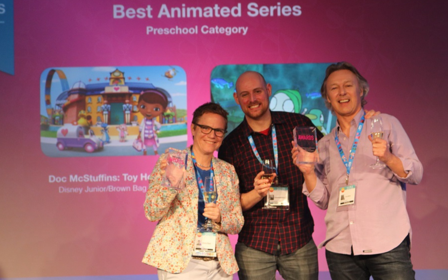 Three people on stage holding awards and champagne glasses in front of a screen reading “Best Animated Series – Preschool Category.”