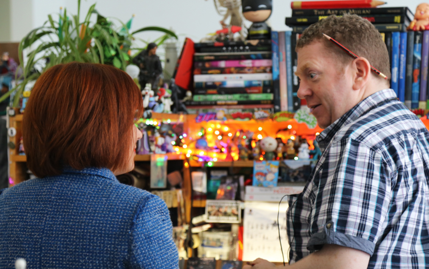 Two people talking in a workspace decorated with books, figurines, and colorful lights.