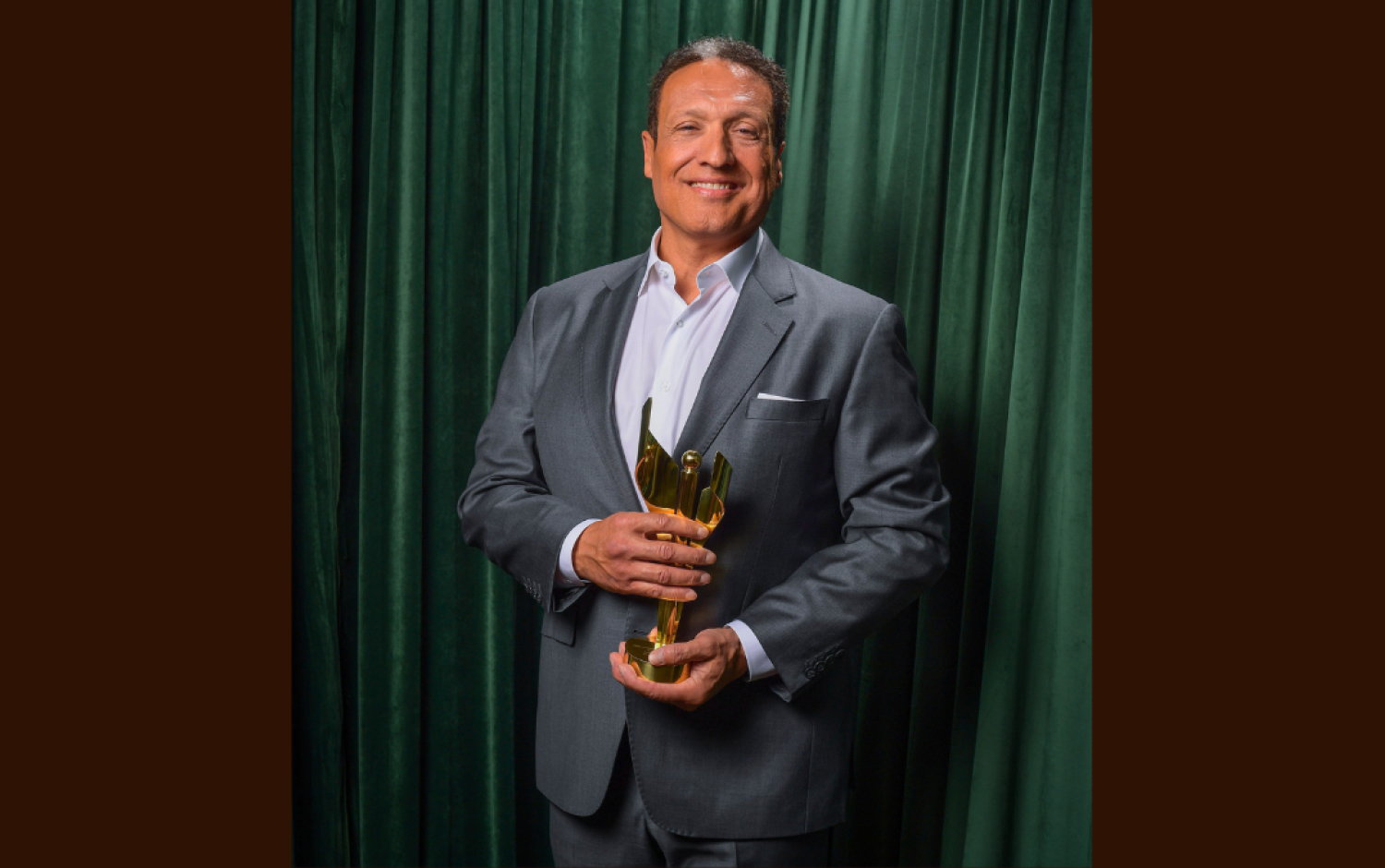 Person in a gray suit holding a golden award trophy against a green curtain backdrop.