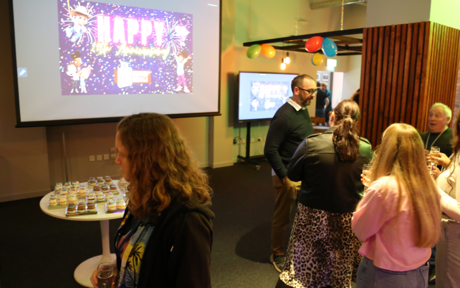 People mingling near a table of cupcakes in a room with a “Happy Anniversary” screen and colorful balloons.