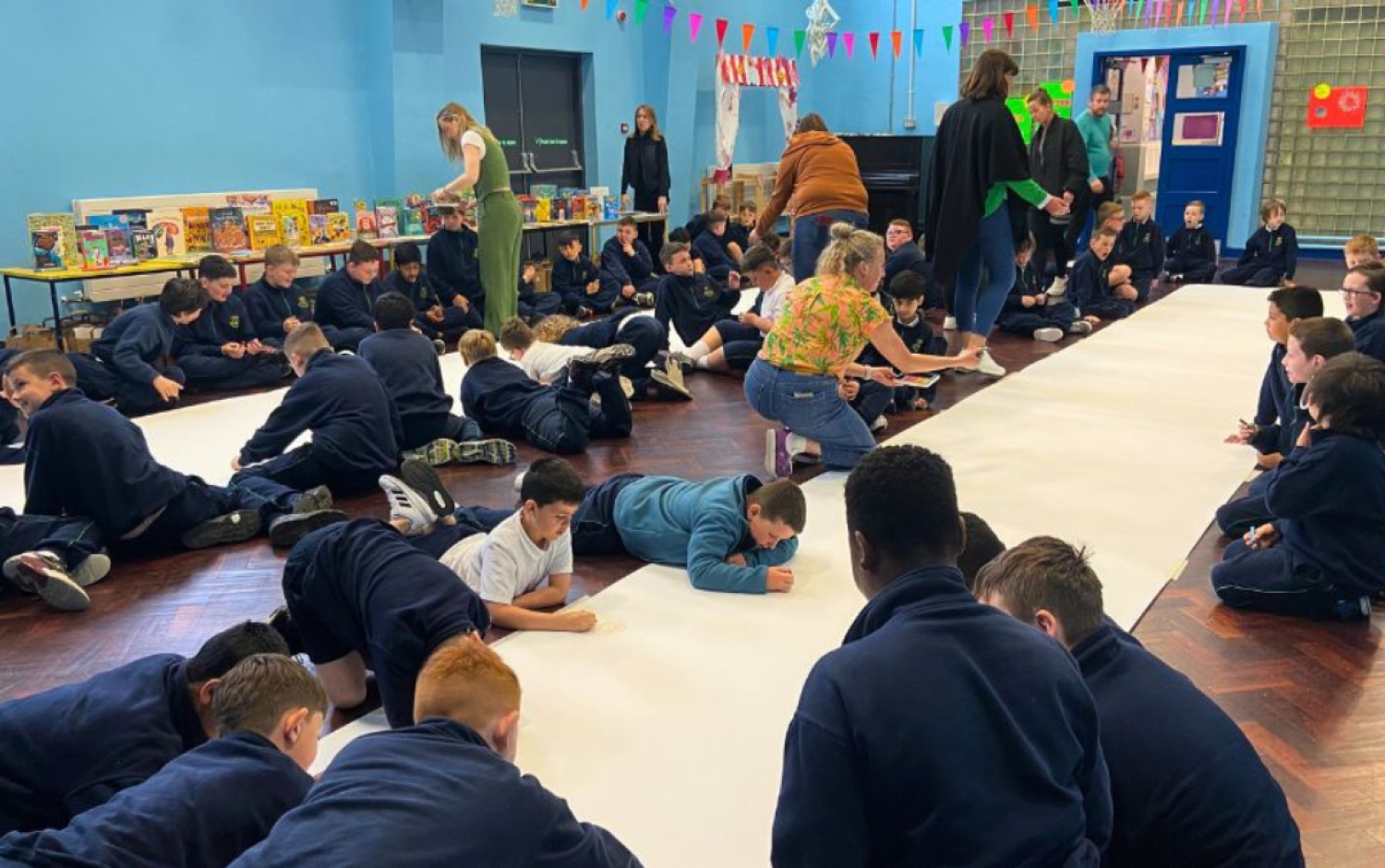 Large group of students lying on the floor drawing on long sheets of paper during an art activity.