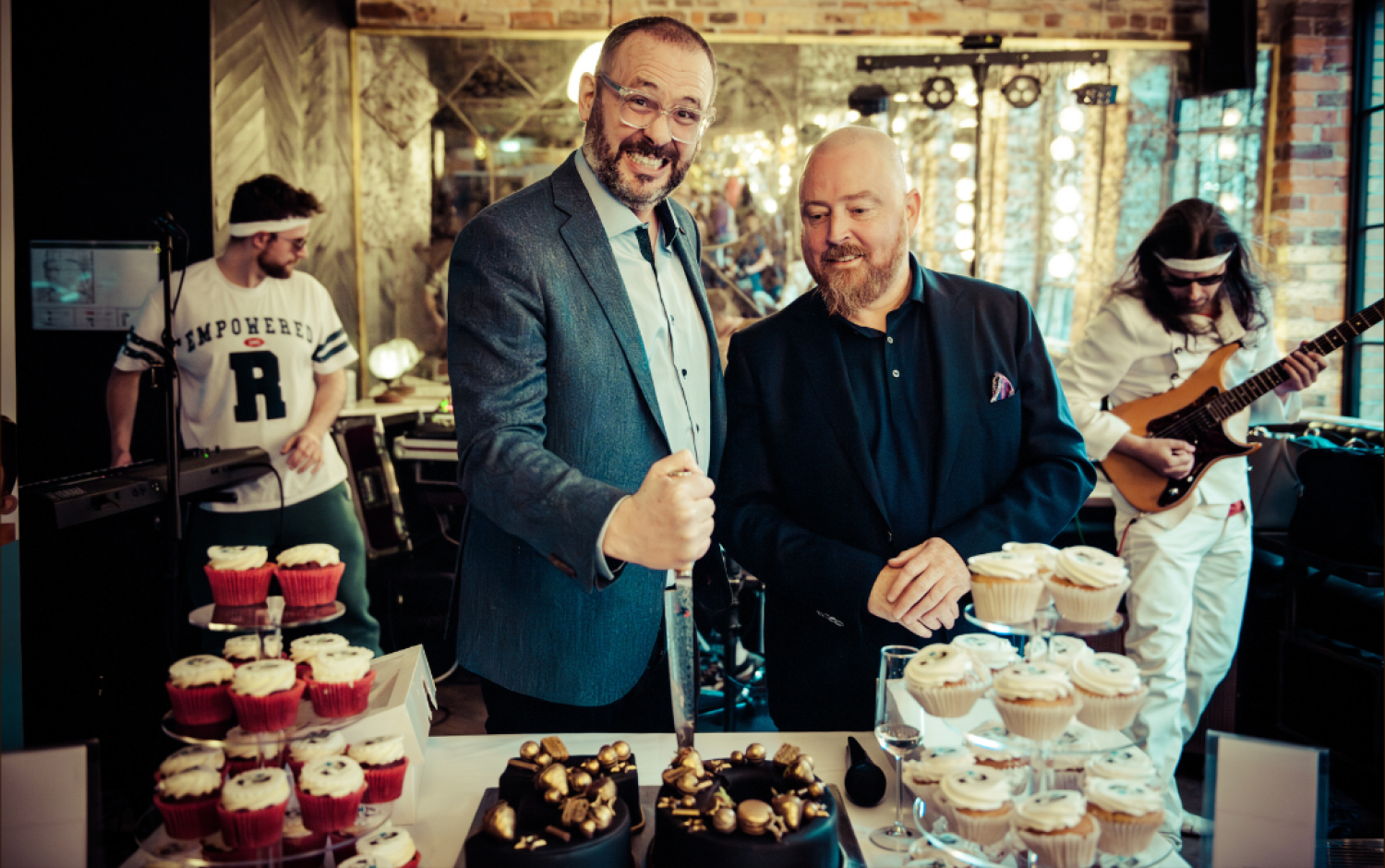 Two people standing behind a dessert table with cakes and cupcakes, musician playing guitar in the background.